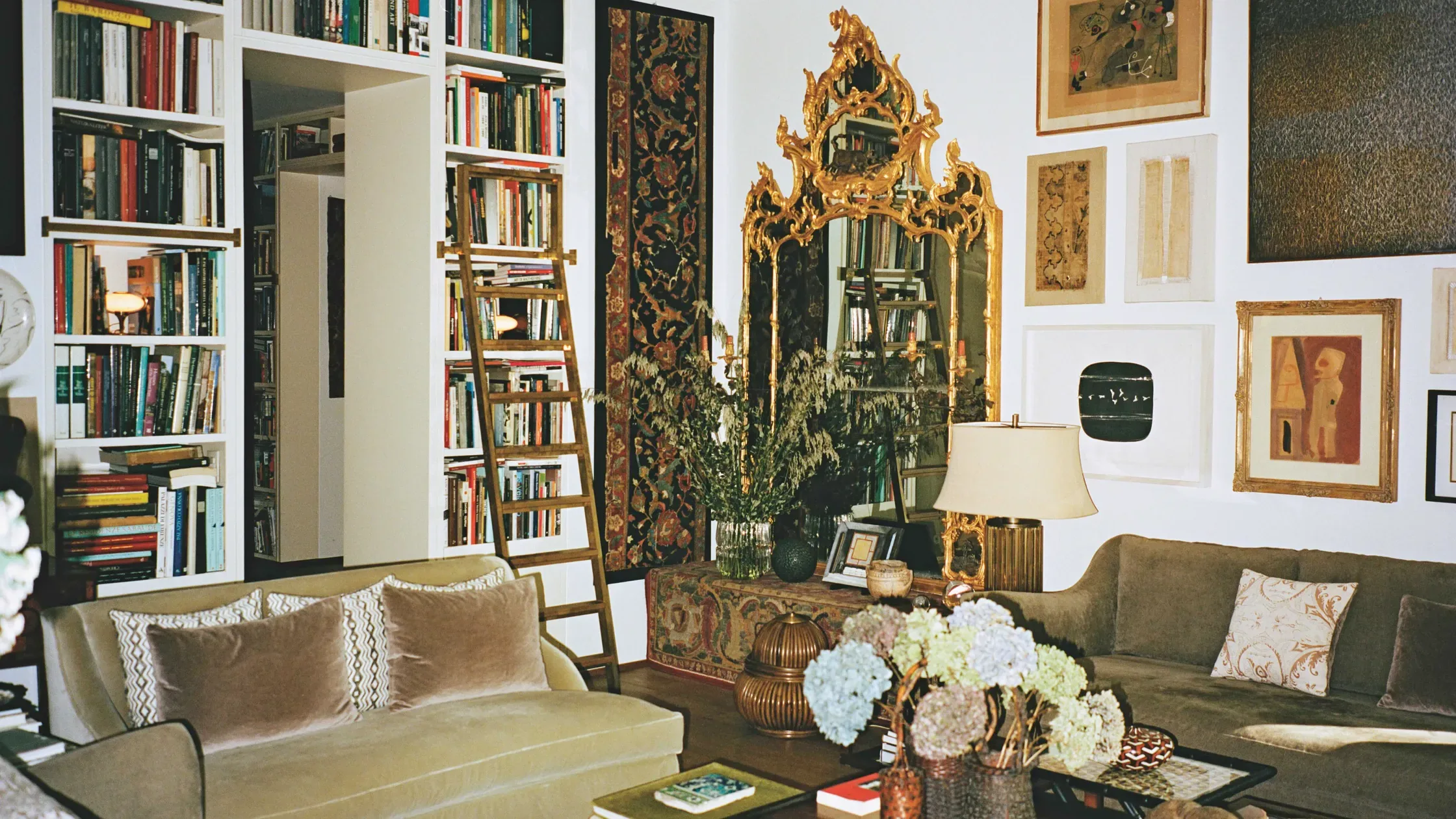 A living room framed by extensive bookshelves, centred around an ornate gilt mirror. Sofas, flowers, textiles and framed artworks fill the space, creating a dense yet organised interior.