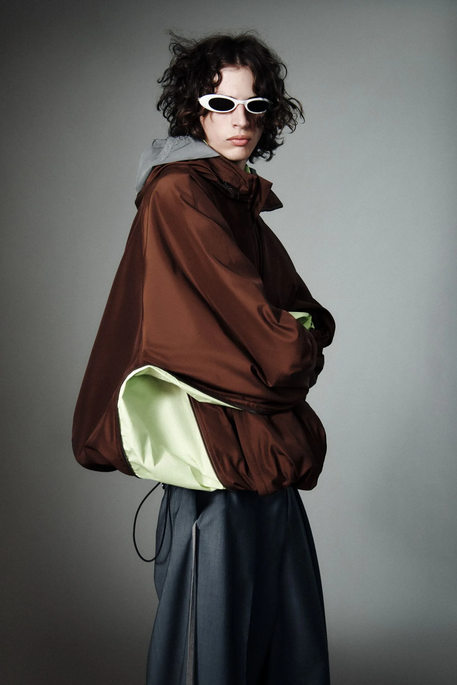 Model in oversized brown jacket, pale green layer, grey skirt and white sunglasses.
