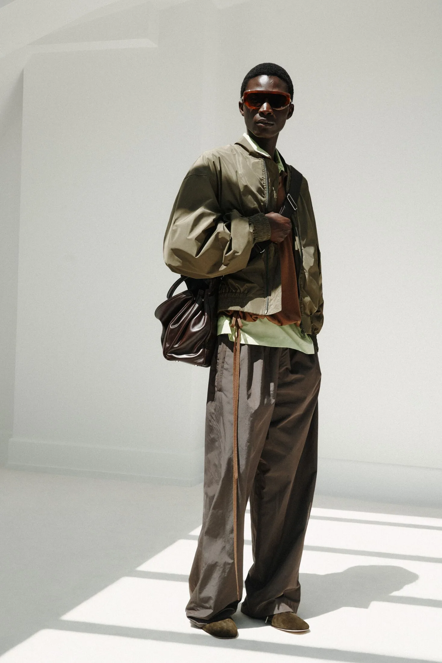Model in khaki jacket, brown trousers, sunglasses and shoulder bag.